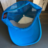 Neon blue and white trucker hat with classic logo, mesh back, displayed indoors on a rustic wooden surface.