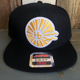 Black 5-panel snapback hat with sunburst design on front, fabric material, resting on a wooden surface.