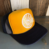Navy, gold, and navy trucker hat with a white logo, displayed on a wooden surface. Mesh back offers breathability.