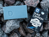 Blue-gray bar soap labeled BIG ASS BRICK OF MANLY SOAP - MIDNIGHT SWIM sits on gray and brown rocks.